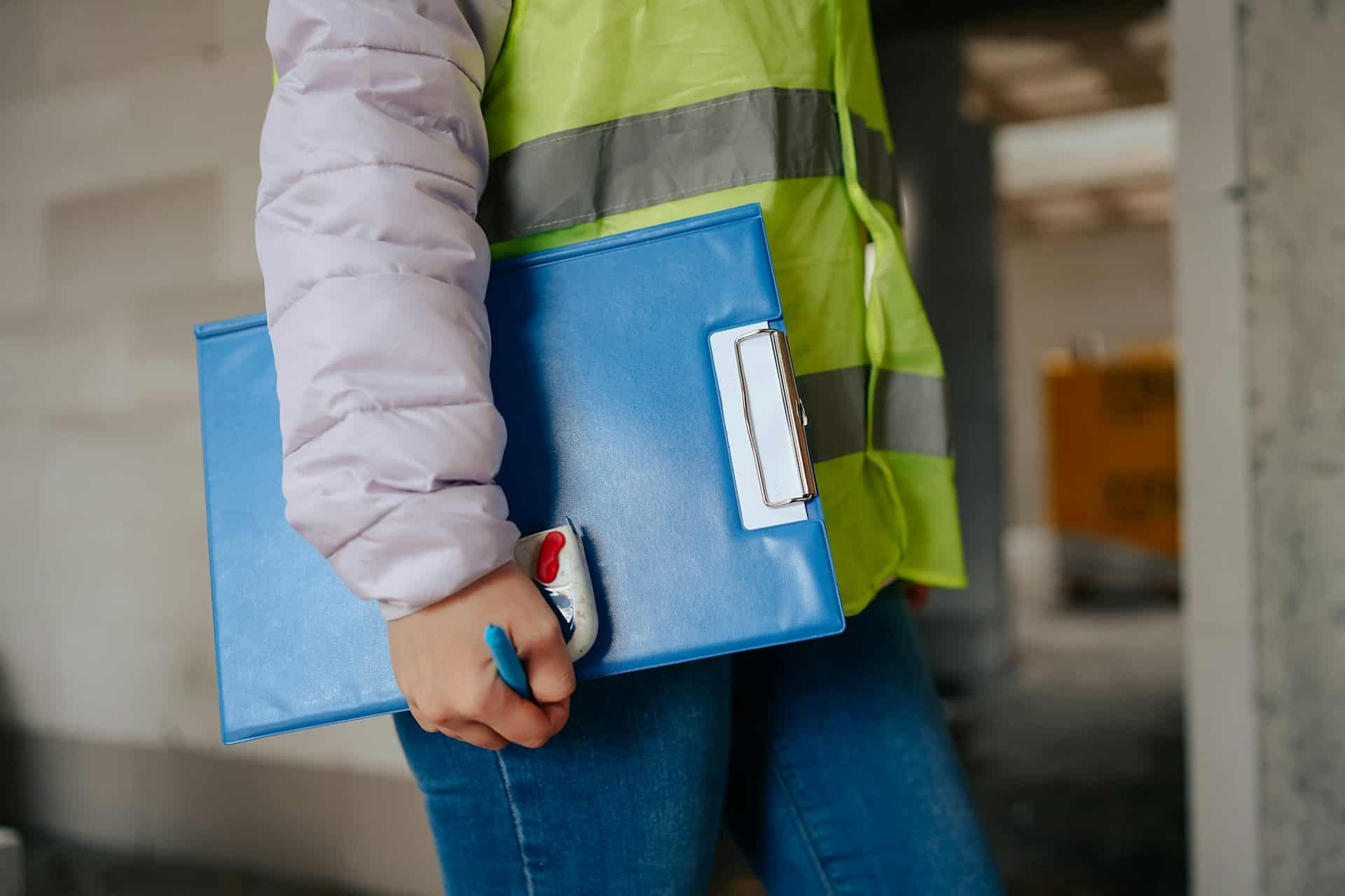 A female builder wearing a high vis vest and holding a blue clipboard.